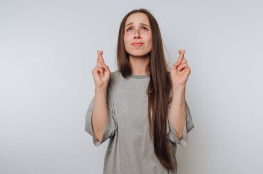 Gri tişörtlü, umut dolu genç bir kadının portresi parmaklarını çapraz yapar, açık arkaplanda bir beklenti hissi oluşturur.