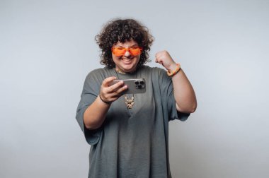 Şaşırmış kıvırcık saçlı, turuncu gözlüklü genç bir kadının portresi açık arkaplanda akıllı telefon kullanırken heyecanla kutluyor.