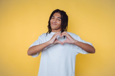 Genç bir adamın portresi elleriyle bir kalp şekli yaratıyor. Sarı arka planda sevgi ve pozitifliği işaret ediyor.