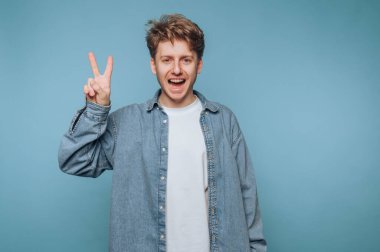 Genç adamın portresi gülümserken bir barış işareti sergiliyor, açık mavi arka planda pozitif enerji yayıyor.