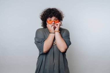 Turuncu güneş gözlüğü takan kıvırcık saçlı genç bir kadının portresi açık arkaplanda şok ve neşe ifade ediyor.