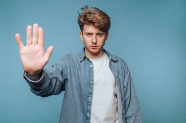 Genç adamın portresi elini kaldırarak açık mavi arka planda dur işareti yapıyor.