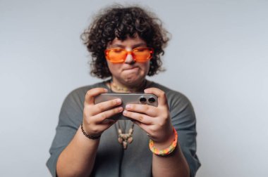 Şaşırmış kıvırcık saçlı genç bir kadının portresi açık arkaplanda akıllı bir telefonla oynarken odaklanmış bir ifade sergiliyor