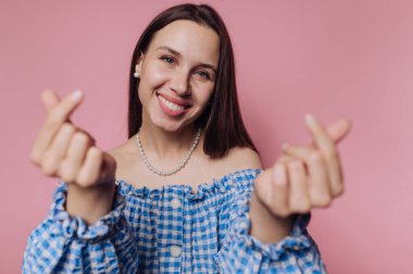 Genç ve gülümseyen bir kadının portresi pembe arka planda mavi kareli bir bluz giyerken elleriyle bir kalp hareketi oluşturur.