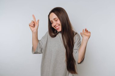 Gri tişörtlü neşeli genç bir kadının portresi açık arkaplanda heyecanla dans ediyor.