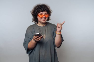 Şaşırmış kıvırcık saçlı genç kadın portresi açık arkaplanda bir telefon tutarken işaret ediyor ve gülümsüyor.