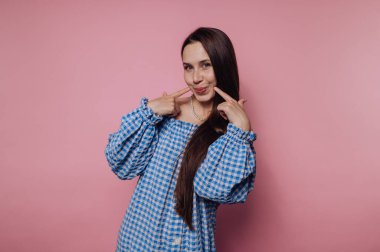 Genç bir kadının portresi, pembe arka planda mavi kareli bir elbise içinde gülüşünü işaret ediyor.