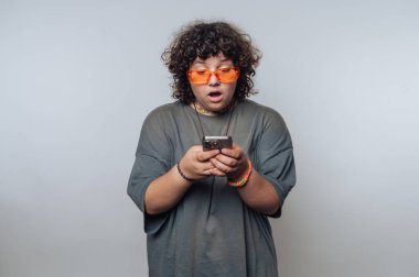 Şaşırmış kıvırcık saçlı genç bir kadının portresi açık arkaplanda akıllı bir telefona bakıyor.