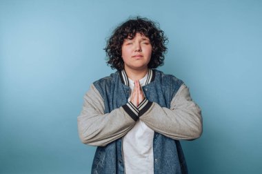 Elleri dua pozisyonunda duran genç bir kadının portresi, mavi arka planda sessizce tasvir edilmiştir.