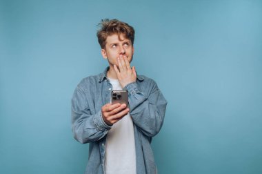 Düşünceli bir gencin portresi elinde akıllı bir telefonla durup açık mavi arka planda yukarı bakıyor.