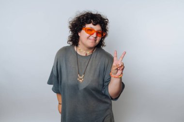 Turuncu güneş gözlüğü takan kıvırcık saçlı genç bir kadının portresi, açık arkaplanda bir barış işaretiyle neşe içinde poz veriyor.