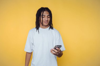 Saçları örgülü genç bir adamın portresi akıllı telefonuna odaklanıyor, sarı arka planda ekrana daldırılmış.