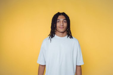 Örgülü saçlı genç adamın portresi kameraya gülümsüyor, sarı arka planda sakin havasını vurguluyor.