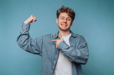 Genç adamın portresi kaslarını kasıyor, açık mavi arka planda güç ve pozitiflik yayıyor.