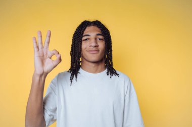 Örgülü genç bir adamın portresi sarı arka planda pozitif ışık saçan bir işaret gösteriyor.