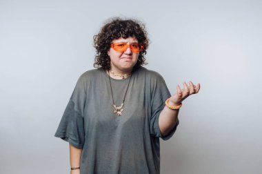 Şaşırmış kıvırcık saçlı, düşünceli, turuncu gözlüklü bir kadının portresi, açık arkaplanda poz verirken bir dizi duyguyu resmediyor.