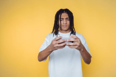 Saçları örgülü genç bir adamın portresi, sarı arka planda oyun makinesiyle uğraşırken endişeli bir görünüm sergiliyor.