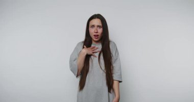 Şaşırmış genç bir kadın ellerini göğsüne dayayıp uzun saçlı ve beyaz arka planda günlük gri tişörtlü şaşkınlığını ifade ediyor.