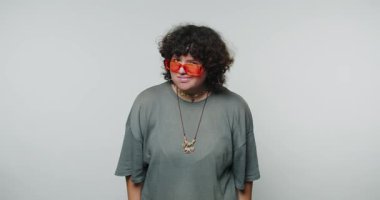 Kıvırcık saçlı, turuncu güneş gözlüğü takan genç bir kadın beyaz arka plandaki rahat kıyafete güveniyor.