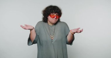 Turuncu güneş gözlüklü genç kıvırcık kadın beyaz arka planda belirsizlik ve neşe ifade ediyor