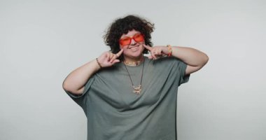 Elleri yanaklarında neşeli, neşeli genç bir kadın pozu veriyor, oyunculuk gösterisi yapıyor ve beyaz arka planda canlı bir kişilik duygusu var.