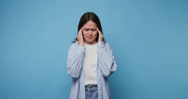 Şakaklarını kederli bir ifadeyle tutan genç bir kız. Mavi arka plandaki rahatsızlığı ya da stresi simgeliyor.