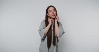 Gülümseyen genç kadın parmaklarıyla yanaklarına hafifçe dokunuyor, beyaz arka planda özen ve şefkat gösteriyor.