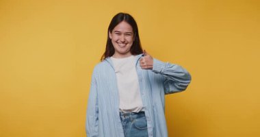 Gülümseyen genç bir kadın olumlu bir jest yapar, pozitif enerji yayar ve hayat dolu sarı arka planda refah verir.