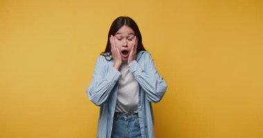 Şaşırmış genç bir kadın, canlı sarı arka planda yüzünü tutarken şok ve şaşkınlık gösteriyor.