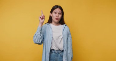 Genç bir kadın parmağını kaldırmış el kol hareketi yapıyor, bir yandan neşeli ve çekici bir tavırla sarı arka planda bir noktayı işaret ediyor.