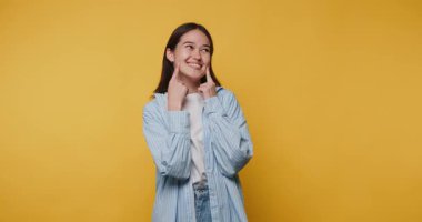 Gülümseyen genç kadın neşeli bir şekilde kendi yanaklarını işaret ediyor, neşeli ve kaprisli bir şekilde sarı arka planda.