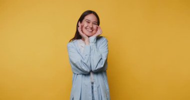 Neşeli genç bir kadın, elleri yanaklarında, parlak sarı arka planda, parlak bir şekilde gülümsüyor.