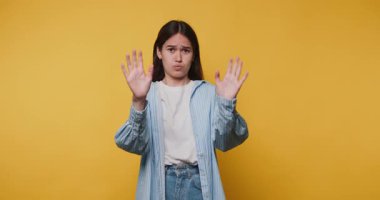 Genç bir kadın, elleri havada, canlı sarı arka planda, tarafsız bir yüz ifadesi sergiliyor.