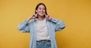 Neşeli insan neşeli bir ifadeyle poz verir, parmakları yanaklarda, mutluluğu yansıtır ve canlı sarı arka planda bağlanır.