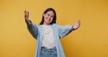 Mutlu genç kadın kollarını açıp jestler yapıyor, sıcaklığı davet ediyor ve ifade ediyor, canlı sarı arka planda bir bağ ve neşe hissi yaratıyor.