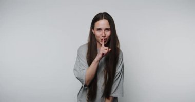 Genç bir kadın şakacı bir şekilde sessiz bir jest yapıyor. Parmağı beyaz arka planda.