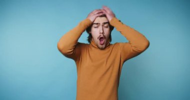 Genç adamın görüntüsü, mavi arka planda dururken kafasını tutarak şoku ifade ediyor.