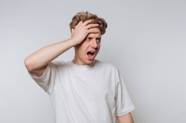 Beyaz tişörtlü genç bir adam şaşırmış görünüyor ve bir eliyle arka planda başını tutuyor..