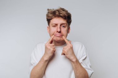 Beyaz gömlekli genç bir adam, parmaklarını arka planda sade bir zemine dayayarak ağzının kenarlarını çekerek komik bir yüz ifadesi takınıyor..