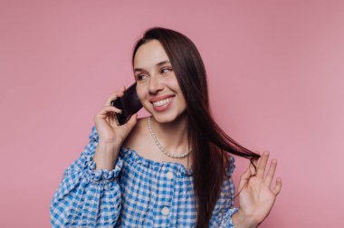 Mavi kareli elbiseli gülümseyen kadın telefonda pembe arka planda konuşuyor..