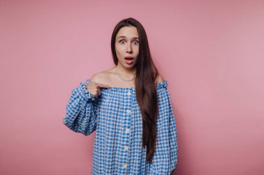 Pembe arka planda, mavi kareli elbiseli, şaşırmış genç bir kadın..