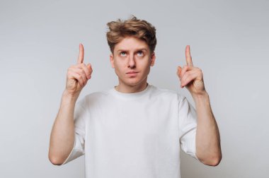 Beyaz tişörtlü genç adam arka planda iki işaret parmağıyla yukarı bakıyor..