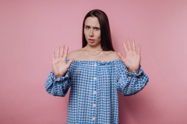 Mavi kareli, omuzları açık bir elbise giymiş bir kadın pembe bir arka plana karşı duruyor, ellerini kaldırıyor ve kendini savunuyor ya da şaşırıyor..