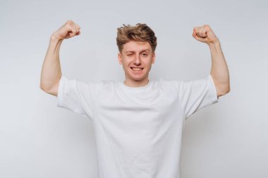 Beyaz tişörtlü genç adam arka planda kaslarını esnetiyor ve göz kırpıyor..