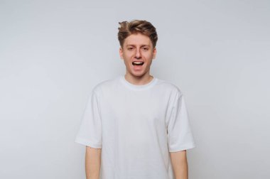 Beyaz tişörtlü genç adam arka planda dikiliyor, kameraya açık ağızla bakıyor, muhtemelen konuşuyor ya da şarkı söylüyor..