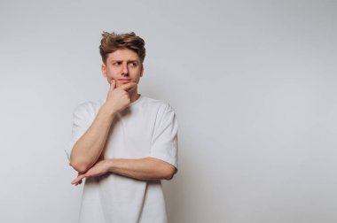 Beyaz tişörtlü genç bir adam düz bir arkaplanda duruyor, eli çenesinde düşünceli görünüyor..