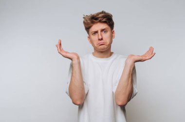 Beyaz tişörtlü genç adam arka planda karışık bir ifadeyle omuz silkiyor..