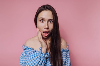 Pembe arka planda, mavi kareli bluzlu, uzun saçlı, şaşırmış bir kadın..