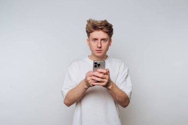 Şaşırmış bir suratı olan genç bir adam, beyaz bir arka plana karşı akıllı telefon tutuyor..