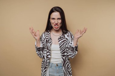Uzun siyah saçlı, zebra desenli gömlek ve kot pantolon giyen bir kadın bej arka planda duruyor, elleri havada ve ağzı açık bir şekilde öfkesini ifade ediyor..